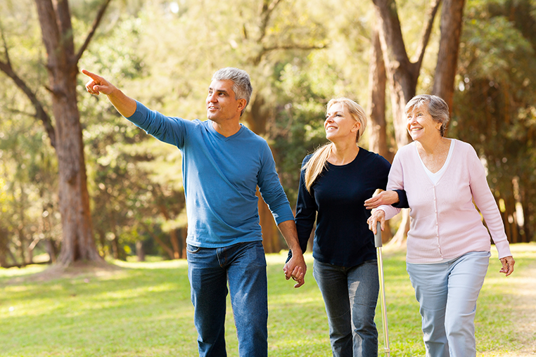Three people walk together through a grassy, wooded area. A man and a woman hold hands, and the woman links arms with an older woman with a cane.